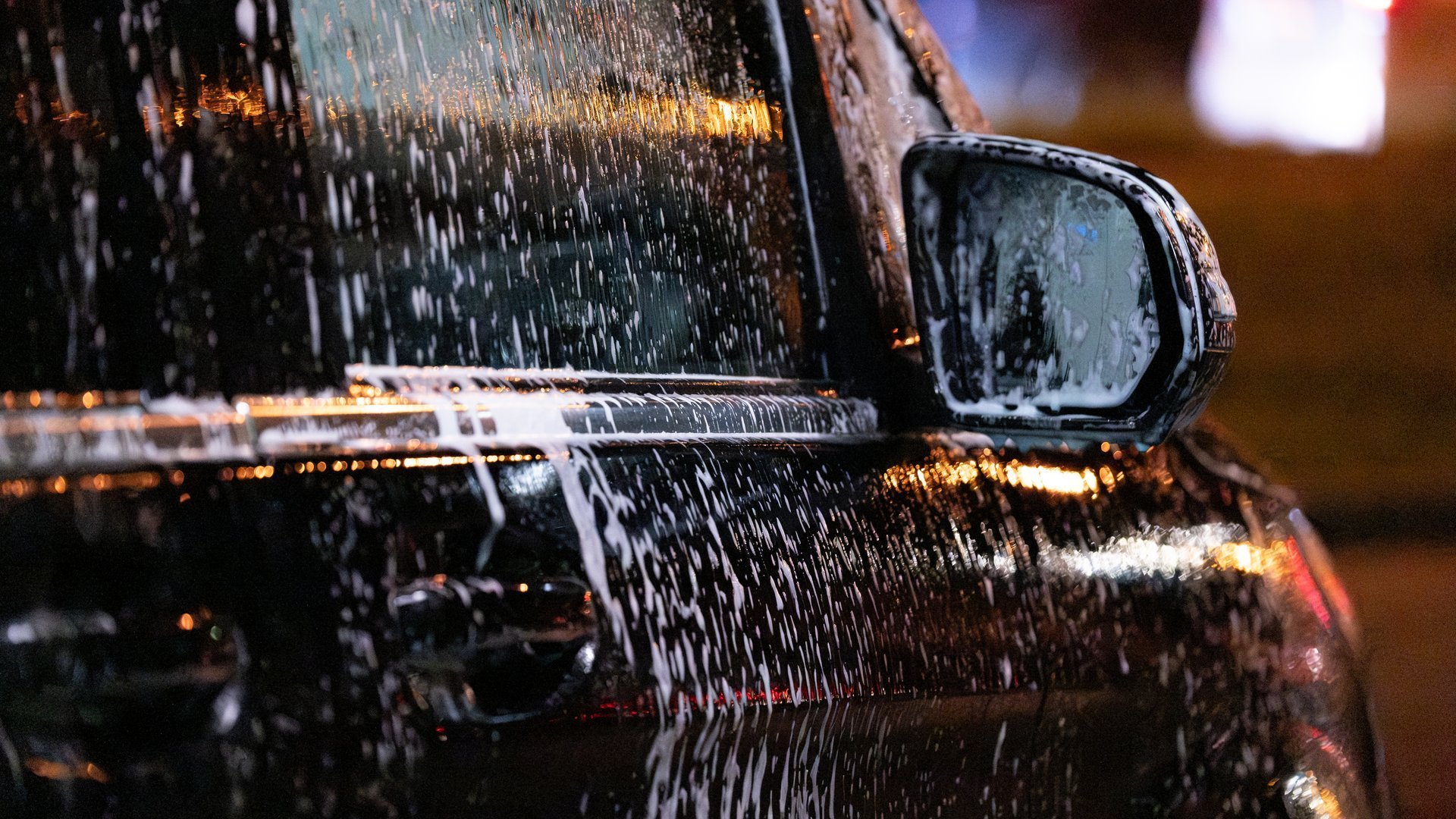 Close-up of a black car covered in soap and water during washing at night. Reflections of city lights highlight the glossy surface and foam texture.